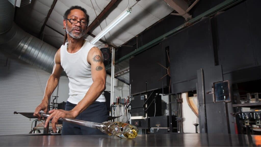 Black man working with glassblowing tools in a studio setting.