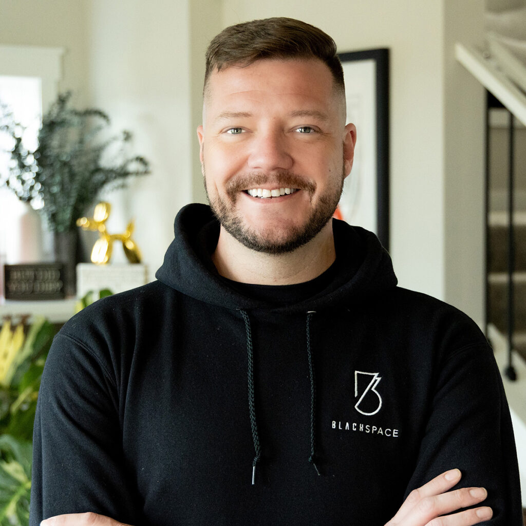 BlackSpace logo hoodie, smiling man in modern home interior, about BlackSpace community and culture.