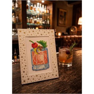 Cross-stitch art of a cocktail glass with a cherry and pineapple on a bar table.